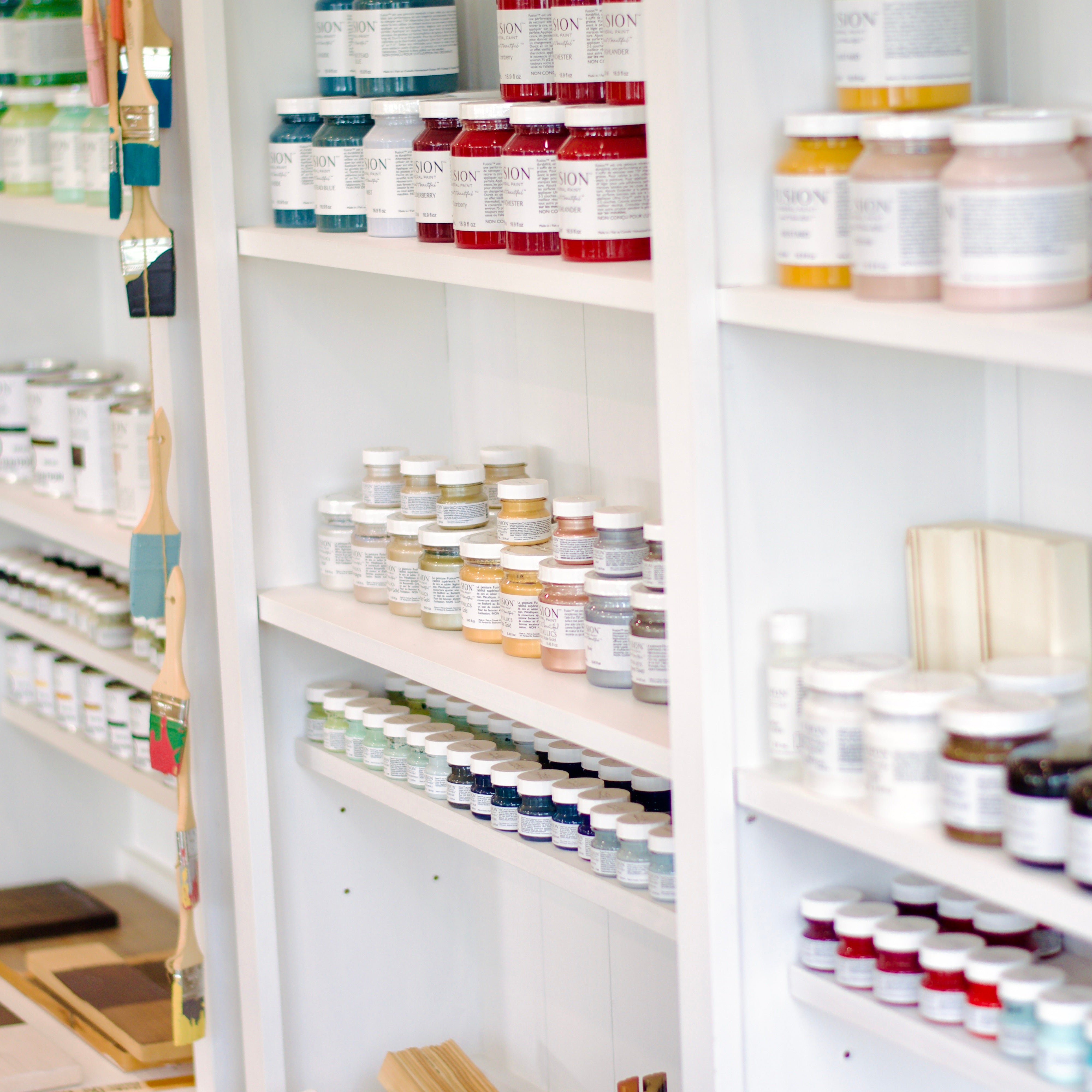 Shelves displaying a variety of Fusion Mineral Paint jars organized by color, with paintbrushes hanging beside them.