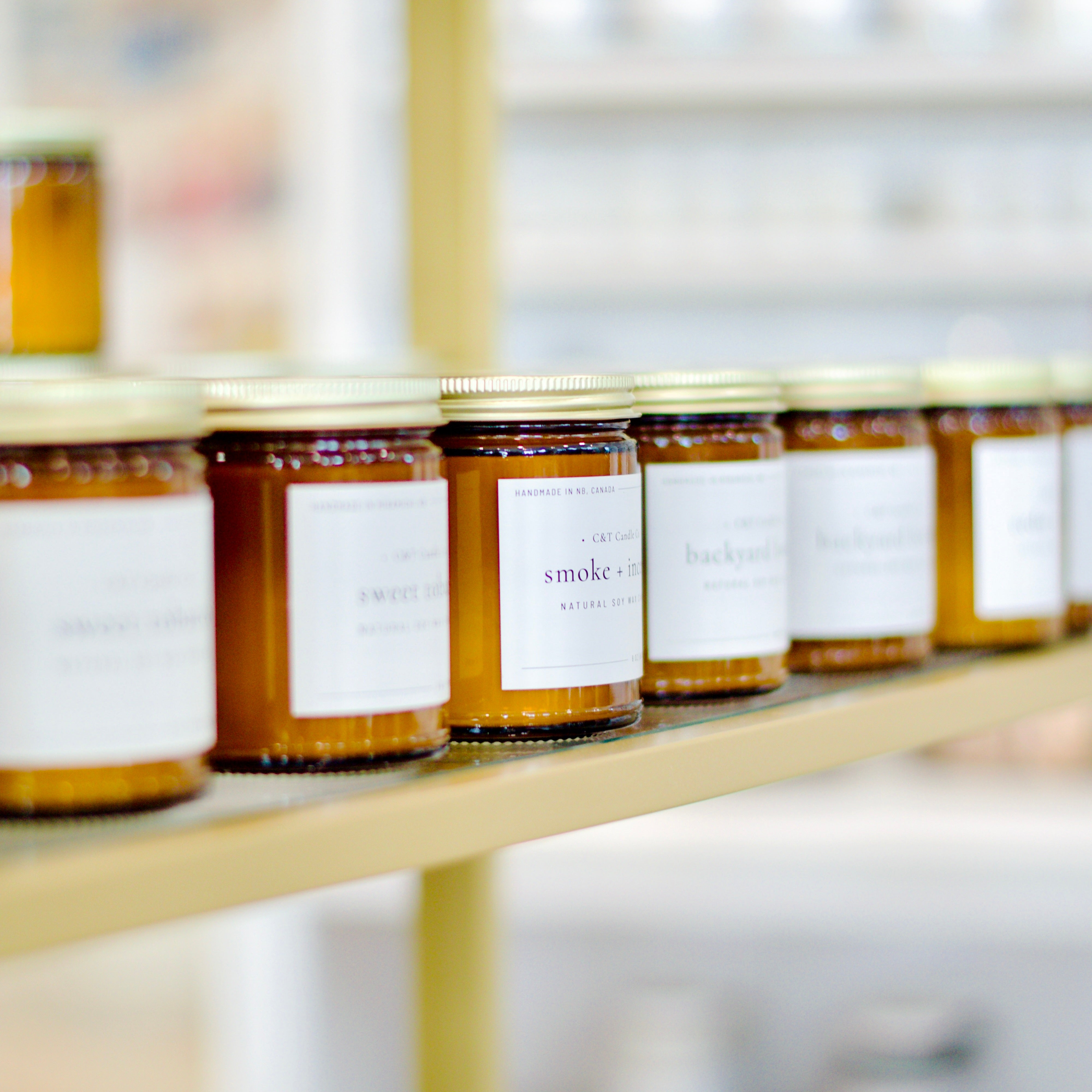 Close-up of a row of amber glass jars with gold lids, labeled with different scents, including 'Smoke + Incense,' displayed on a shelf.