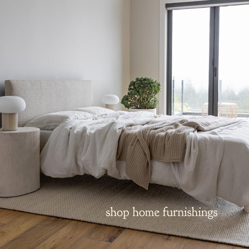 Modern bedroom with a neutral-toned bed featuring soft white and beige linens, a textured upholstered headboard, and wooden bedside tables with round lamps. A large window in the background shows an outdoor view with greenery and a woven accent stool. Overlay text reads 